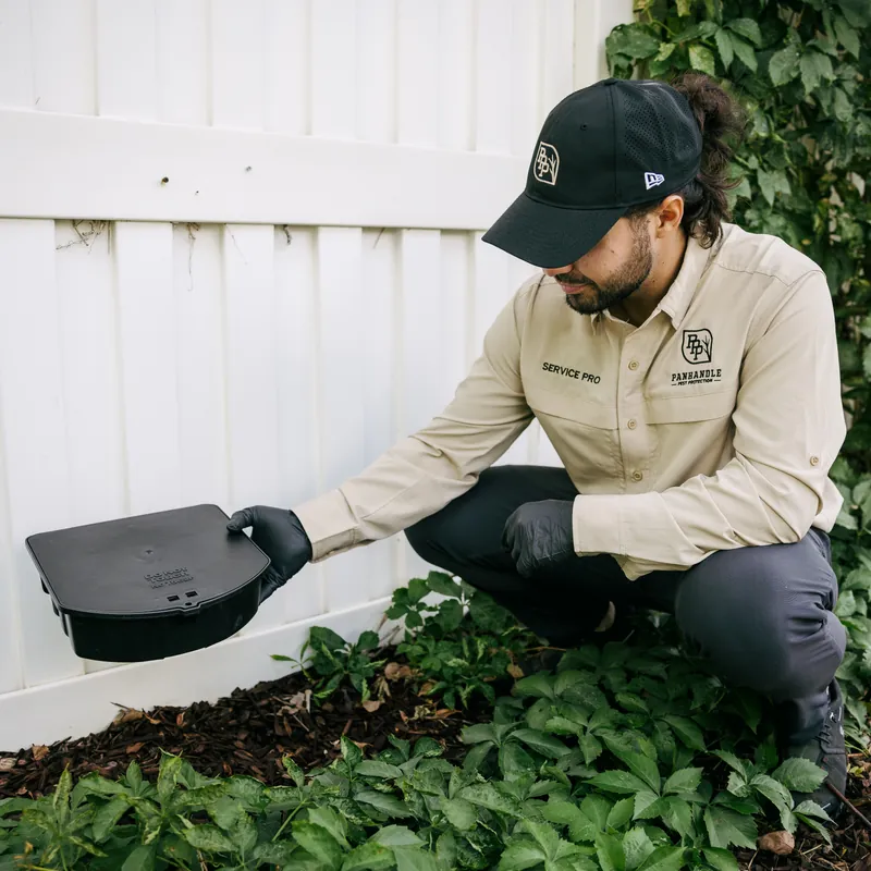 Técnico de control de plagas de Panhandle Pest Protection colocando una estación de cebo para roedores junto a una cerca