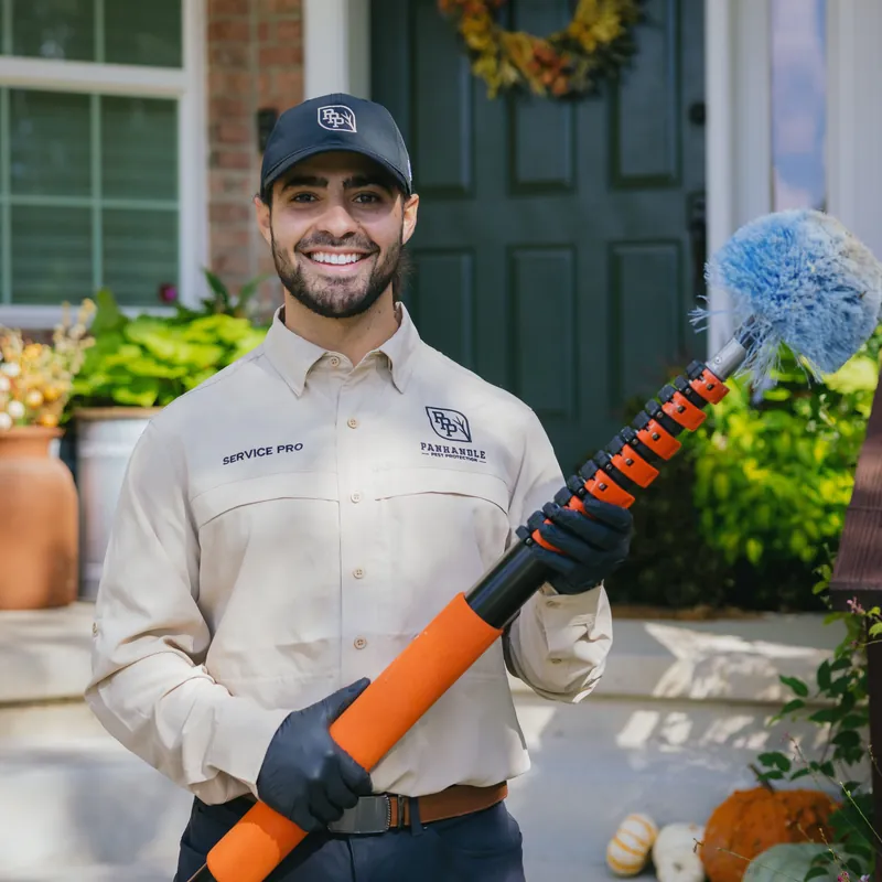 Smiling Panhandle Pest Protection technician with an extendable cobweb duster on a front porch decorated for fall