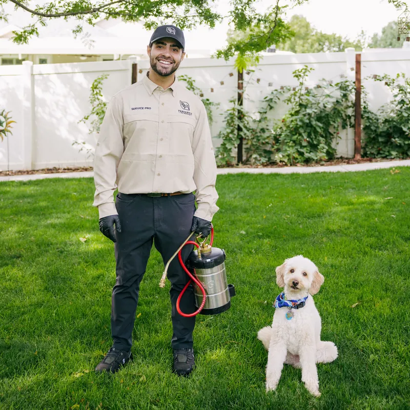 Smiling Panhandle Pest Protection technician with a pump sprayer on a backyard lawn beside a seated dog