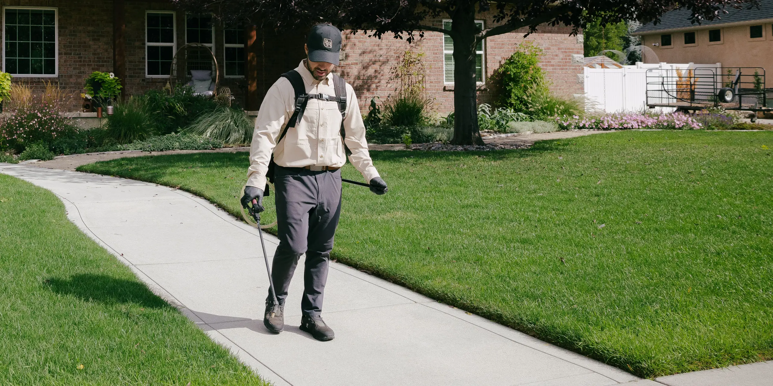 Pest control technician with a backpack sprayer walking a front walkway between green lawns