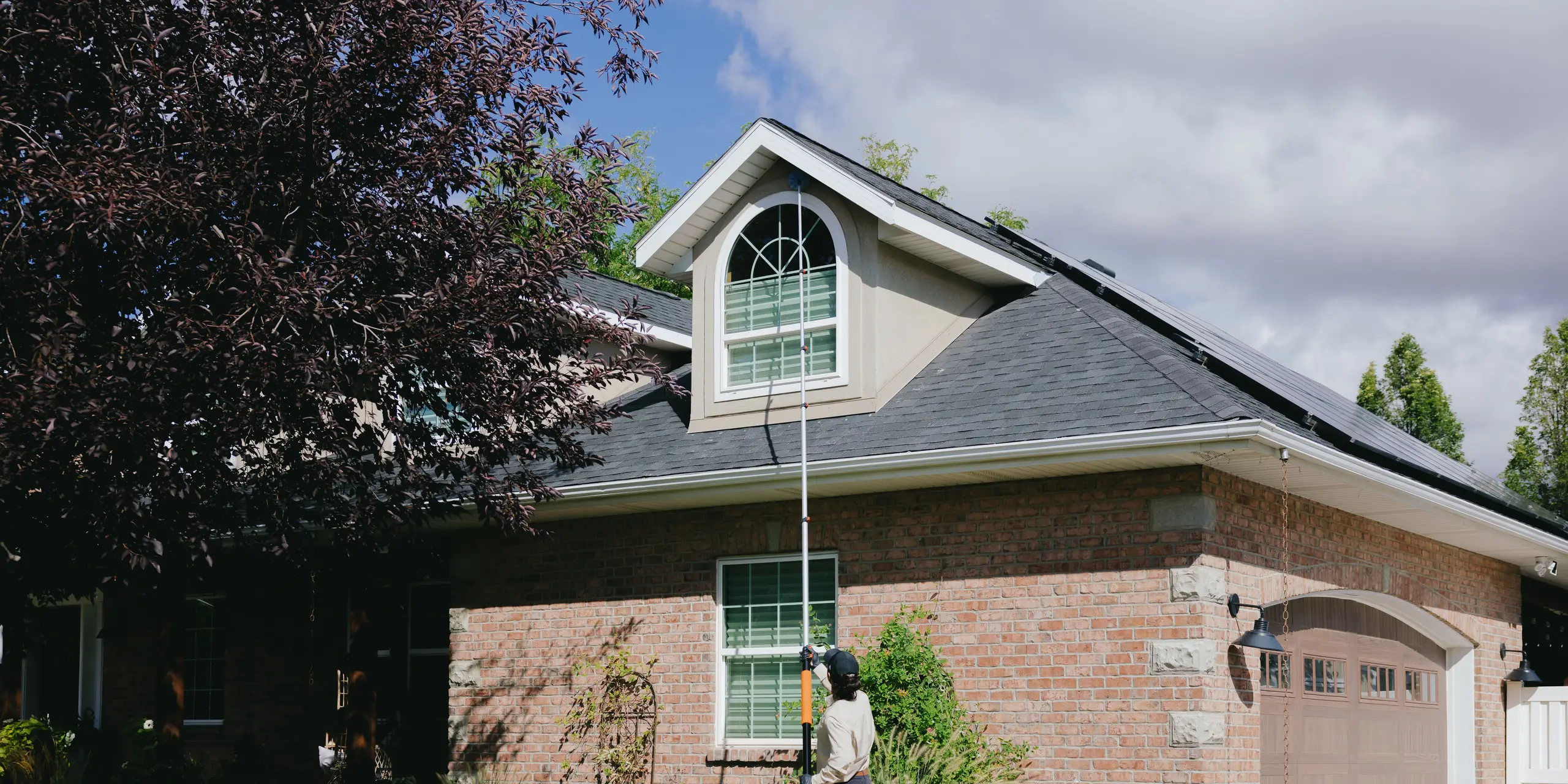 Pest control technician using an extension pole to clear webs from a dormer eave on a brick home