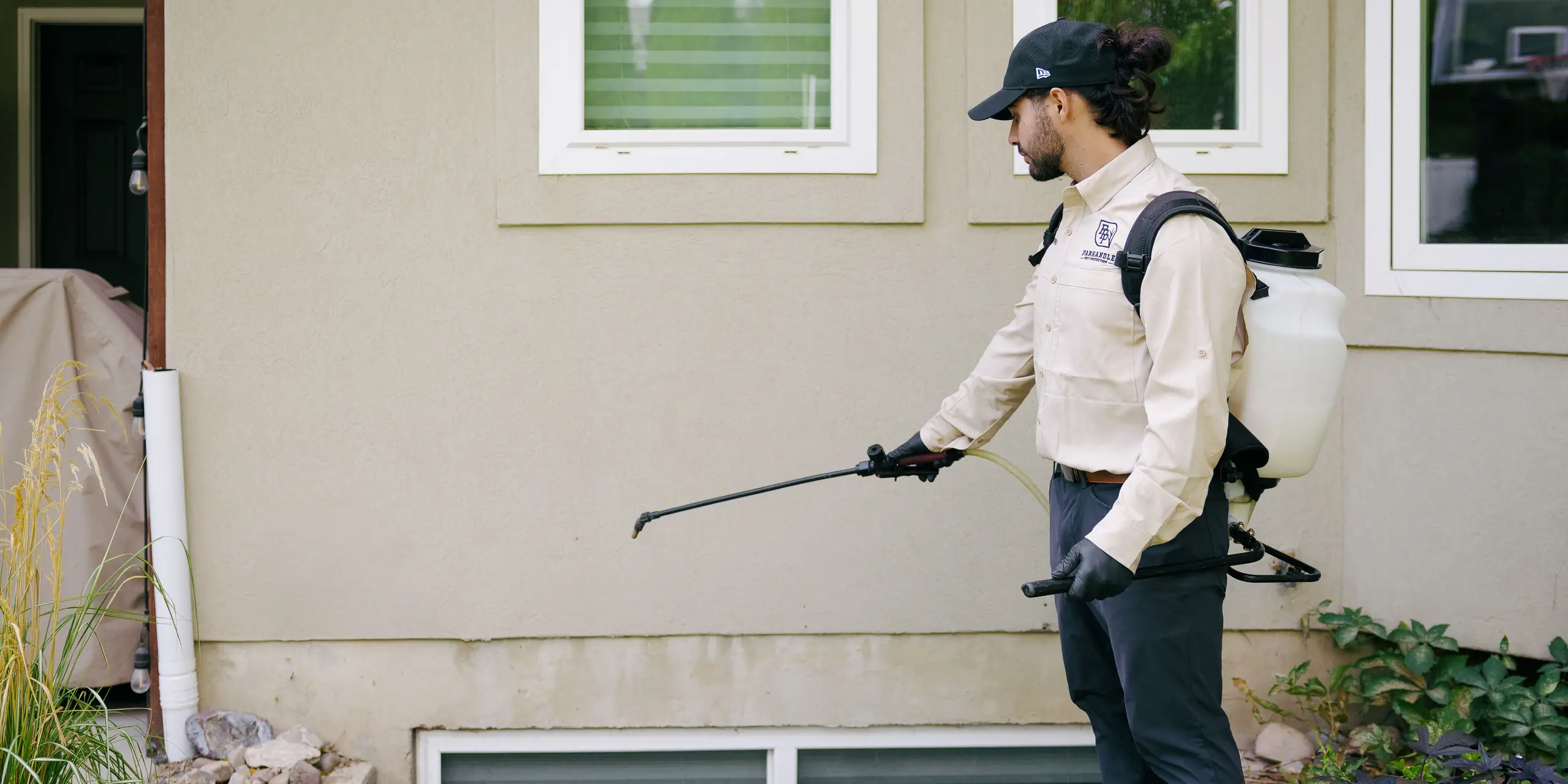 Gloved Panhandle Pest Protection technician treating a home's exterior wall with a backpack sprayer
